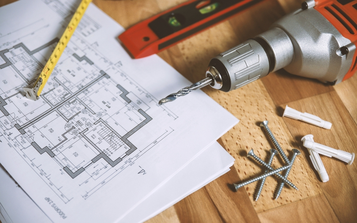 Planos de una vivienda sobre una mesa de madera con taladro, metro y tacos
